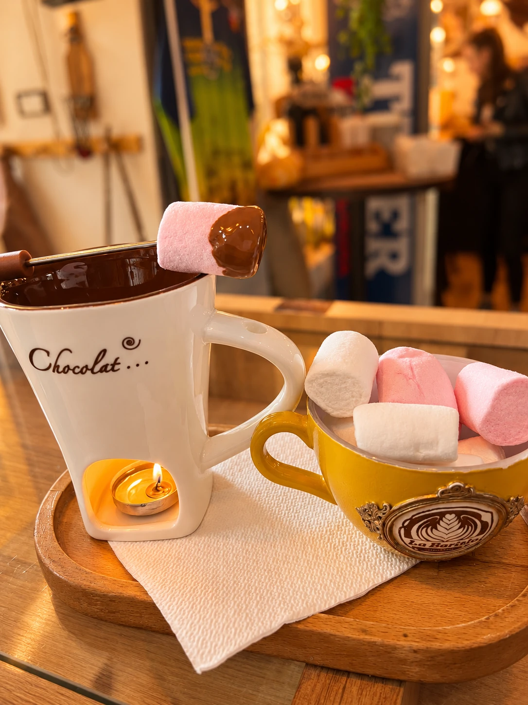 Ciocolată caldă în vasul alb încălzit de lumânare, alături de marshmallows în cana galbenă La Barista
